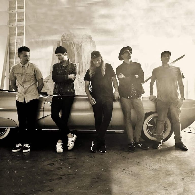 Five band members stand against a vintage car in a monochrome industrial setting.
