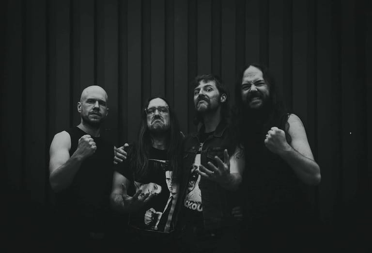 Four musicians in dark clothing perform against a draped stage backdrop in black and white.