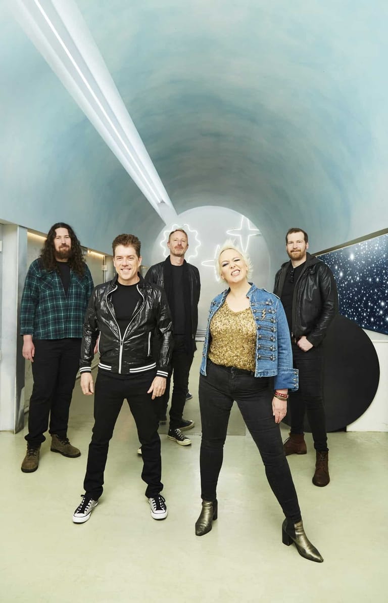 Five musicians stand in a modern tunnel-like venue with white curved walls and dramatic overhead lighting.