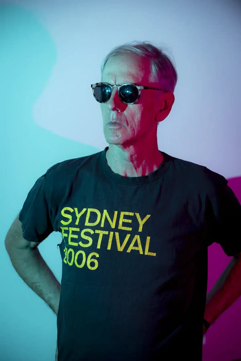 Person wearing dark t-shirt with yellow "Sydney Festival 2006" text and sunglasses, lit by cyan and magenta stage lighting.