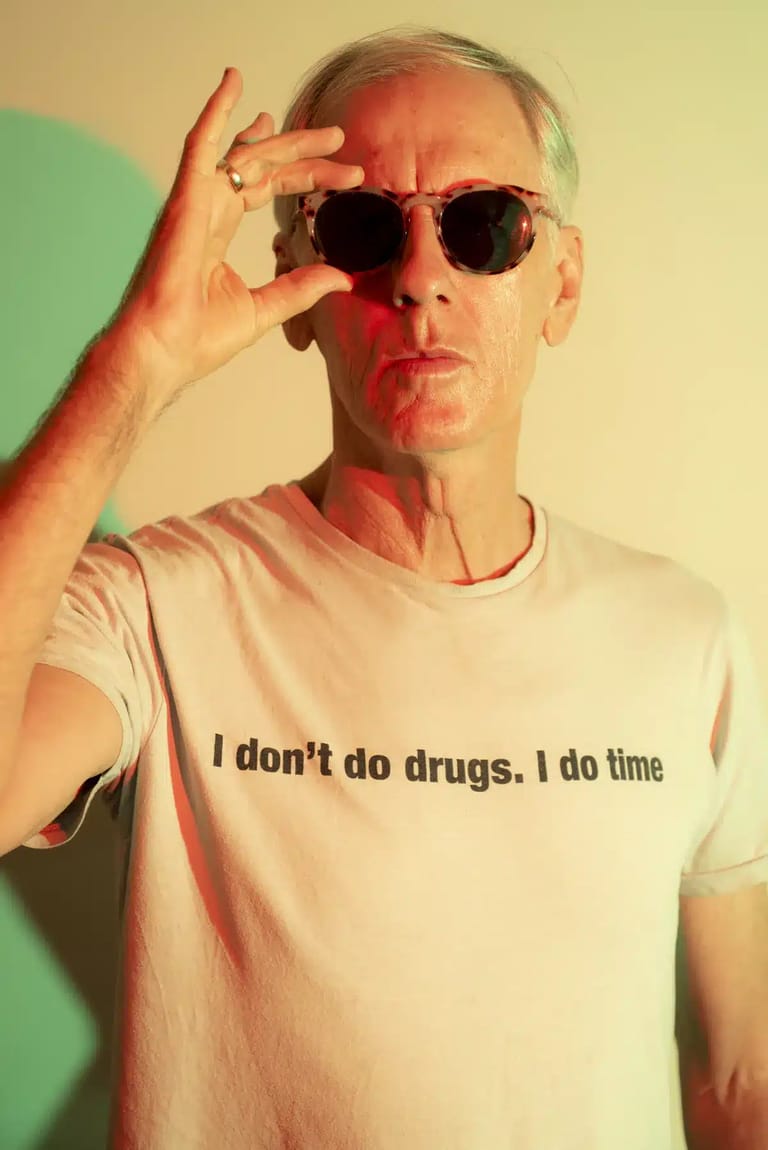 Man wearing sunglasses and text-printed t-shirt adjusting his glasses against a green and yellow backdrop.