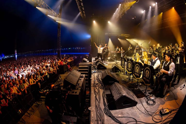 Band performing on stage beneath dramatic golden and blue spotlights before a large crowd in an indoor venue.