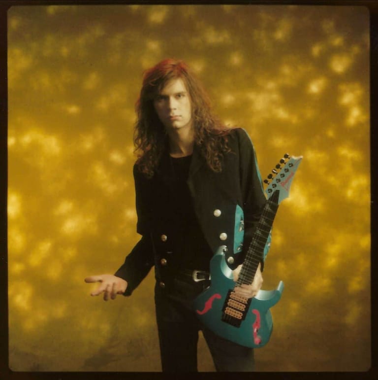 Musician with long reddish hair holding a teal electric guitar against a golden bokeh background.