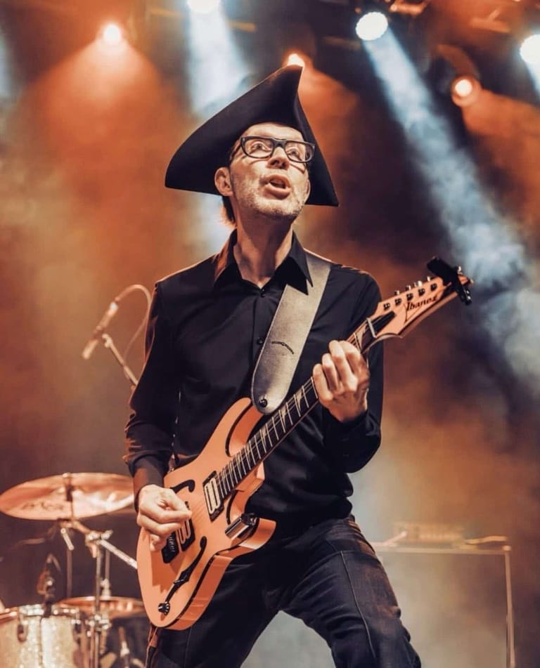 Guitarist in black tricorn hat and glasses performs with electric guitar under warm stage lighting.