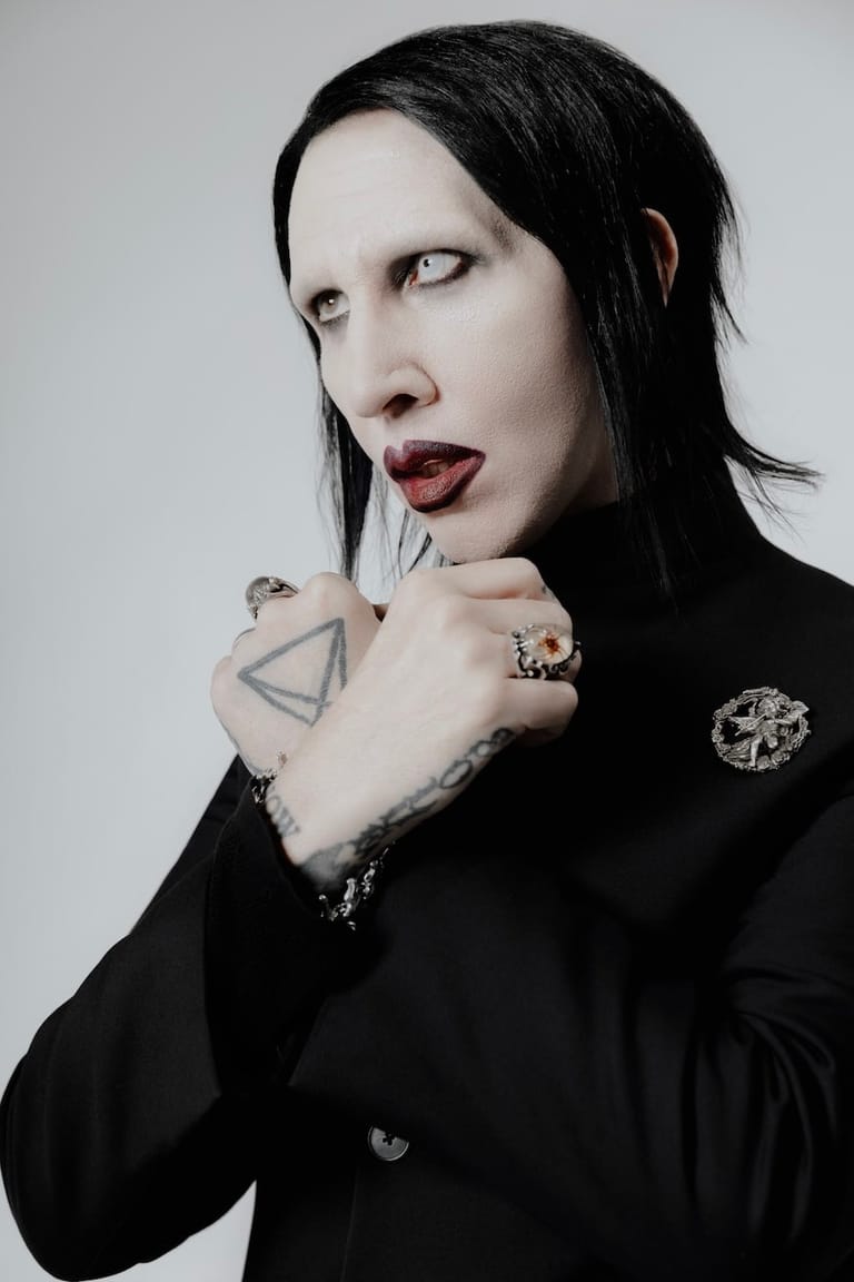 Person with long black hair and dark makeup wearing black clothing, hand raised to face, displaying visible tattoos and rings.