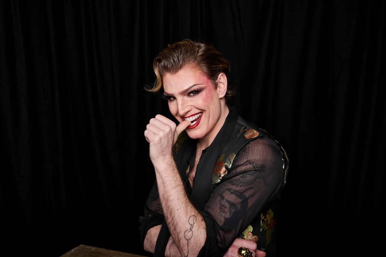 Performer in black patterned shirt smiles at microphone against dark curtain backdrop.