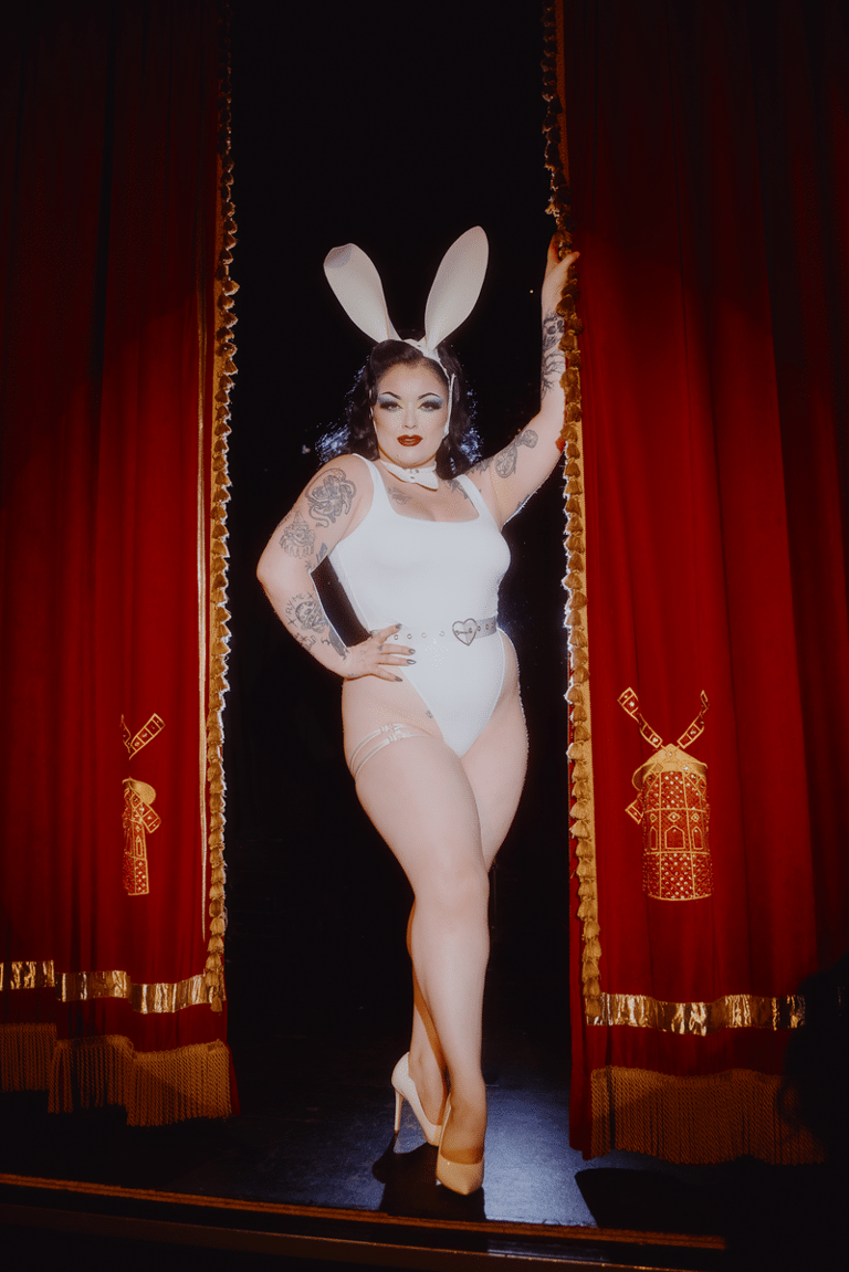 Performer in white bunny costume with oversized ears poses between red velvet curtains trimmed with gold.