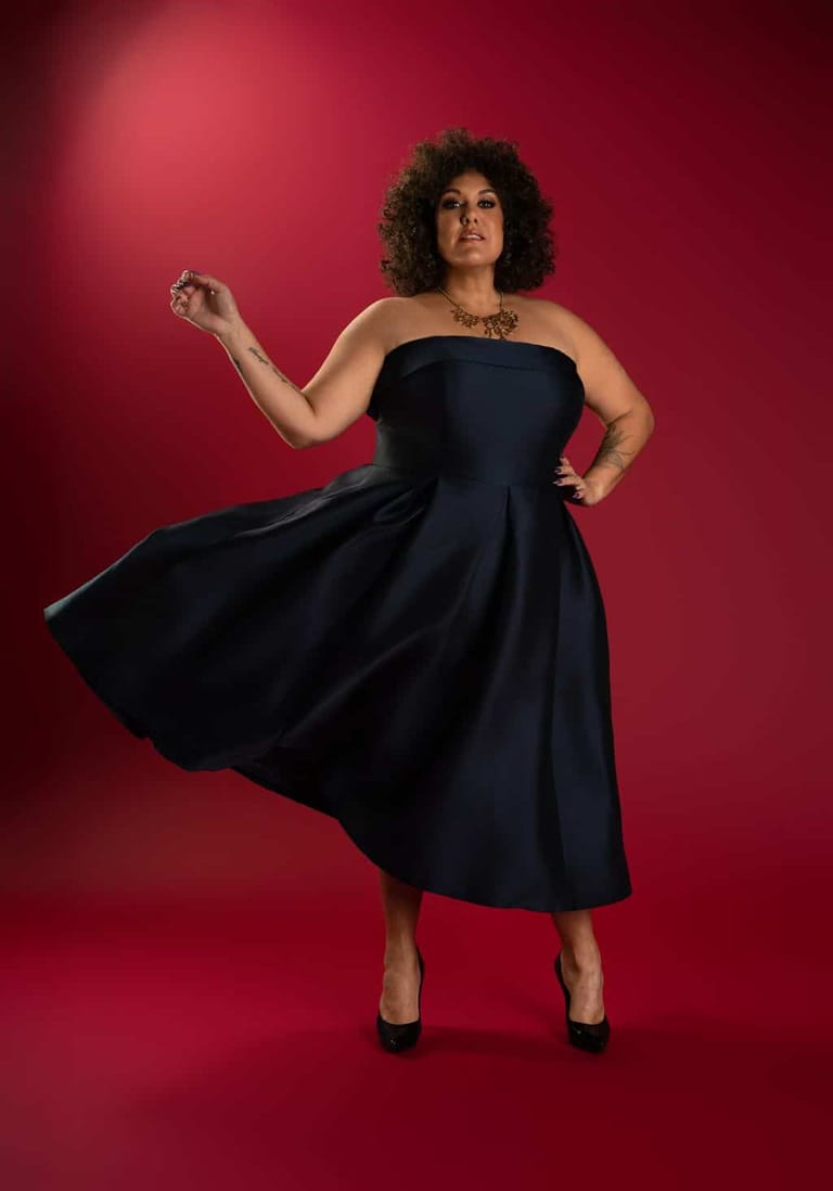Woman in a black strapless dress poses against a vibrant red background, one hand raised thoughtfully.
