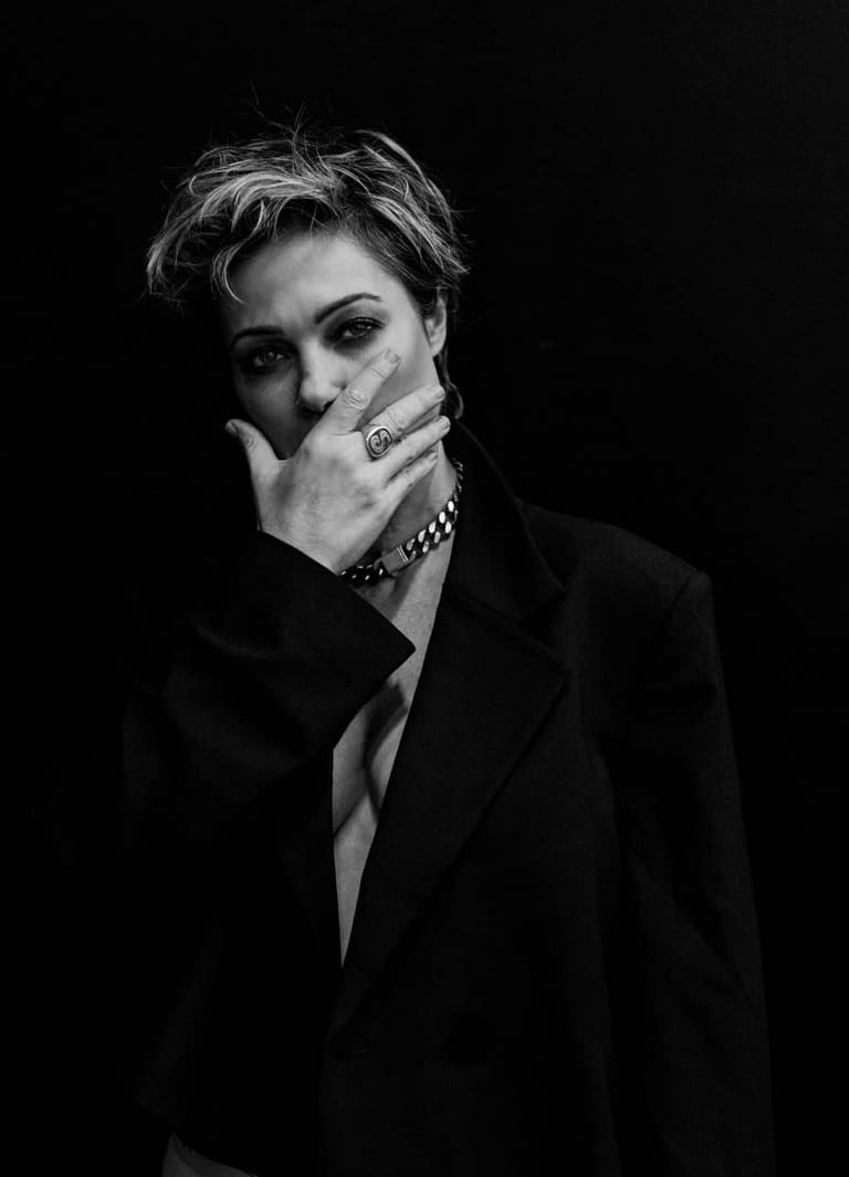 Person with short styled hair wearing a black blazer and chain bracelet, hand raised to face against a dark background.