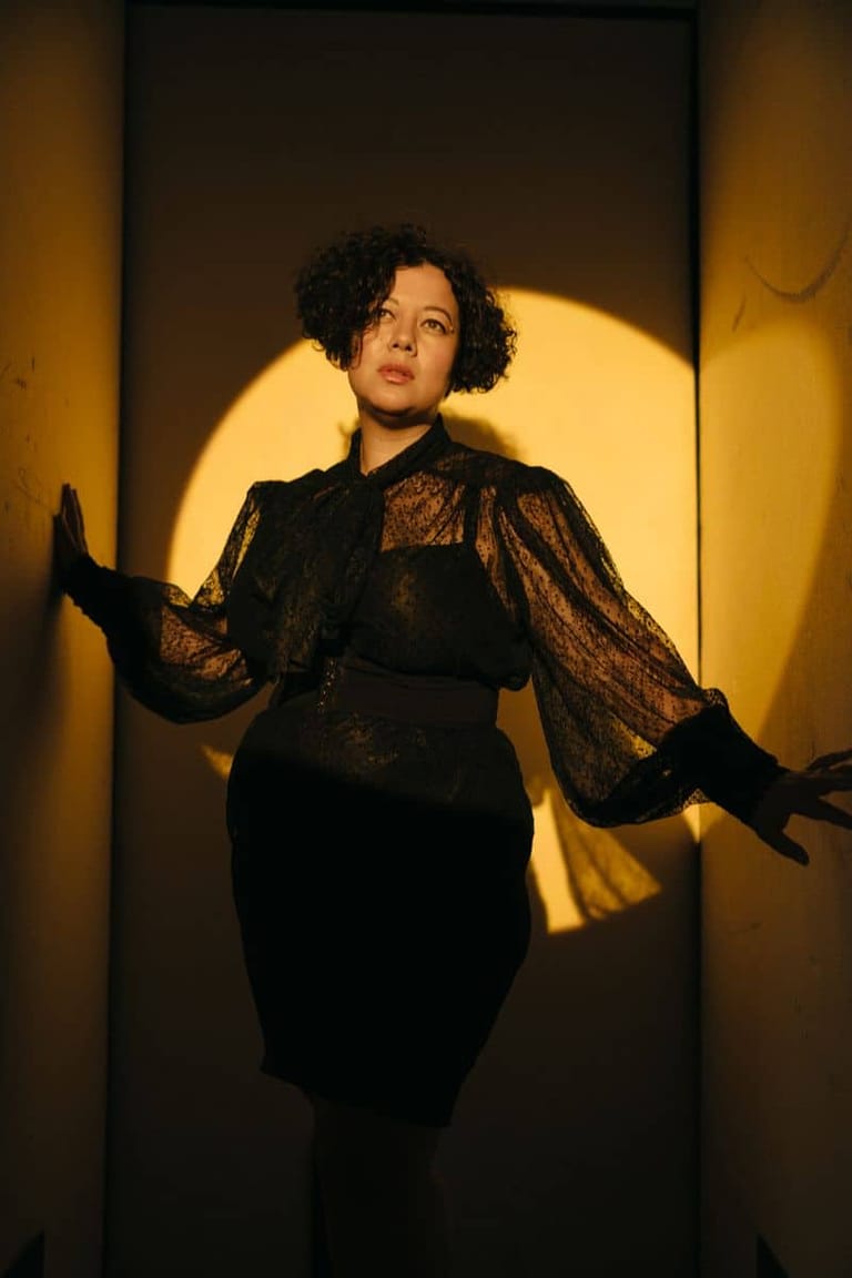 Person in black lace outfit with billowing sleeves posed against warm golden stage lighting.