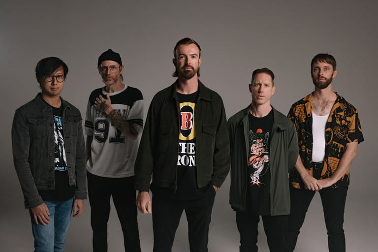 Five band members stand in a line against a neutral grey background during a tour promotion shoot.