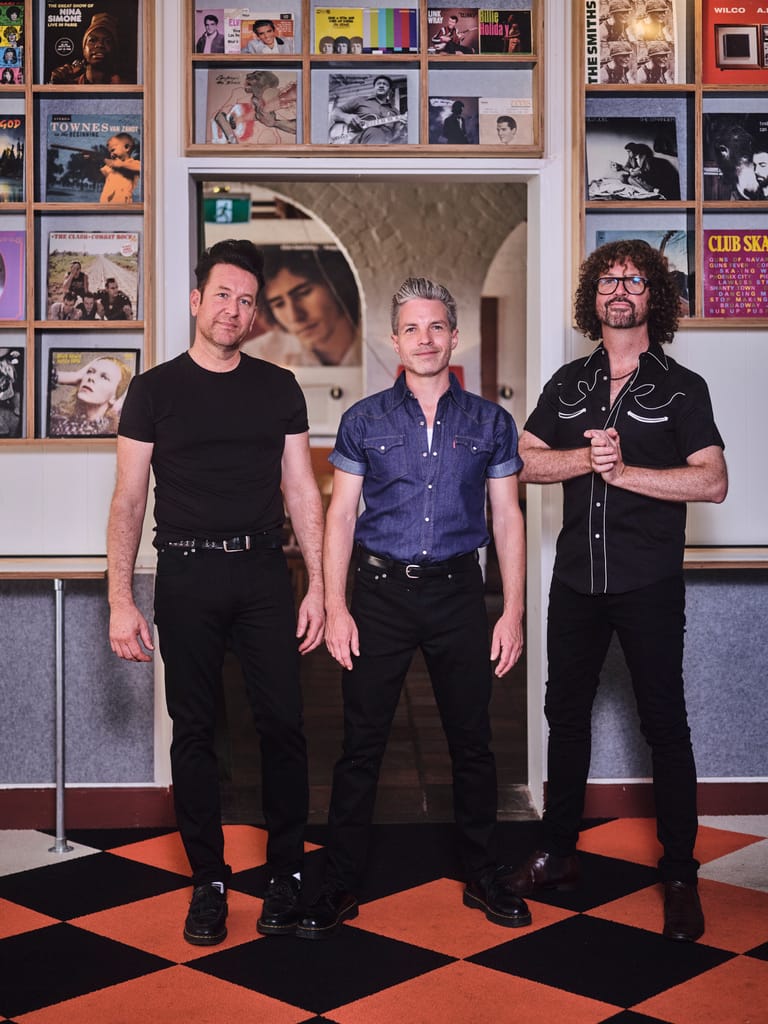 Three band members stand posed in front of a wall lined with vintage music posters and album artwork.