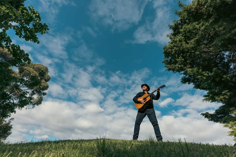 Musician standing in a grassy field playing acoustic guitar against a blue sky with trees framing the scene.