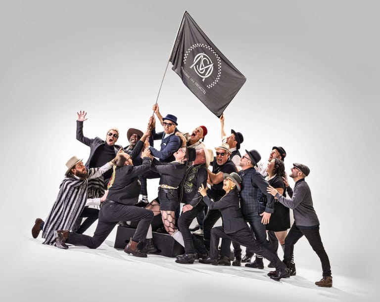 Large group of performers in black outfits clustered together, one holding aloft a dark flag with a circular emblem.