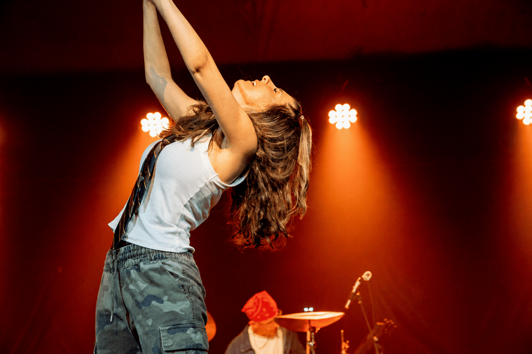 Performer arches backwards on stage with long hair flowing, arms extended upward, beneath warm orange stage lights.