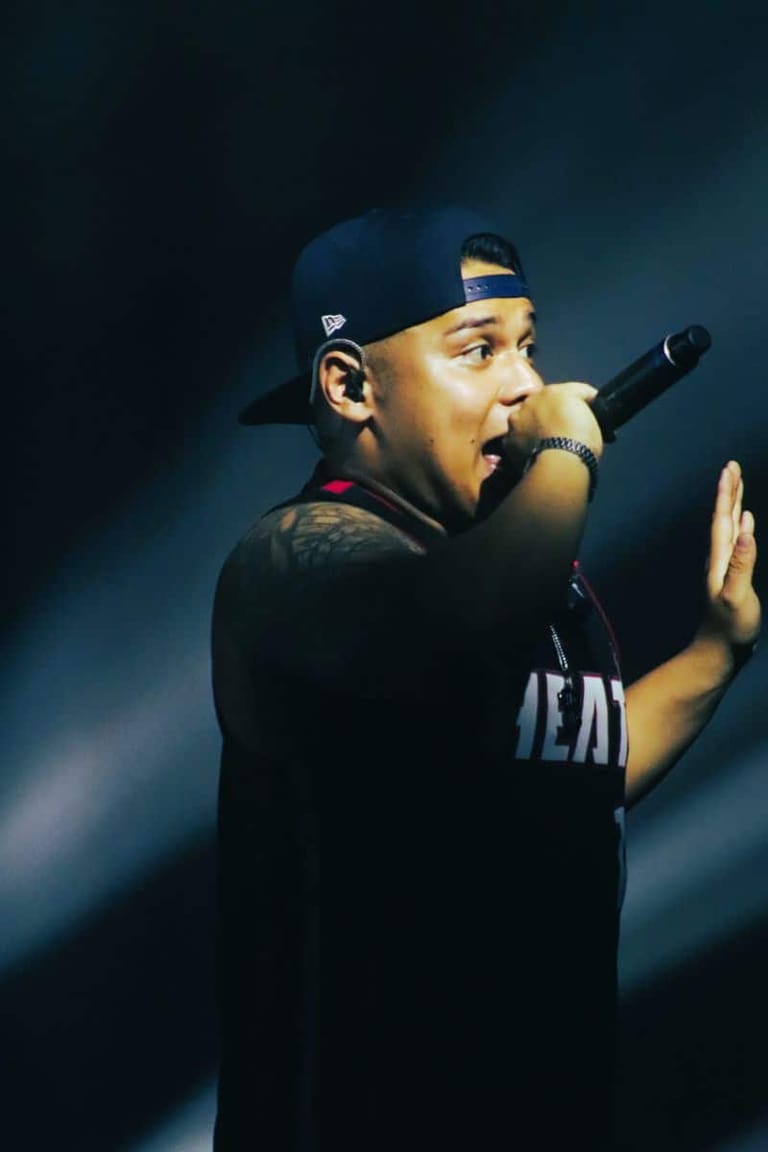Rapper performing energetically at a microphone against a moody blue stage backdrop.