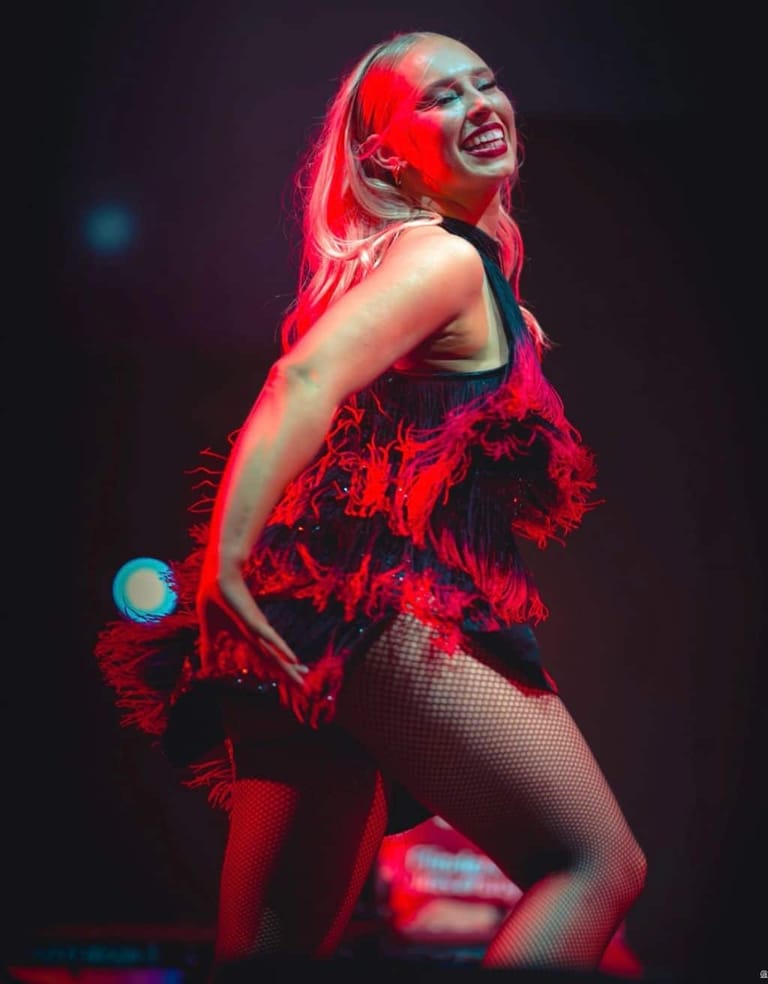 Performer in red feathered outfit and fishnet tights, bathed in red stage lighting during a theatrical production.