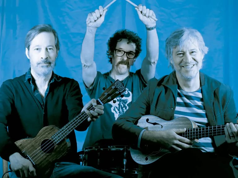Three musicians pose against a blue background, holding bass and acoustic guitars with hands raised above centre figure's head.