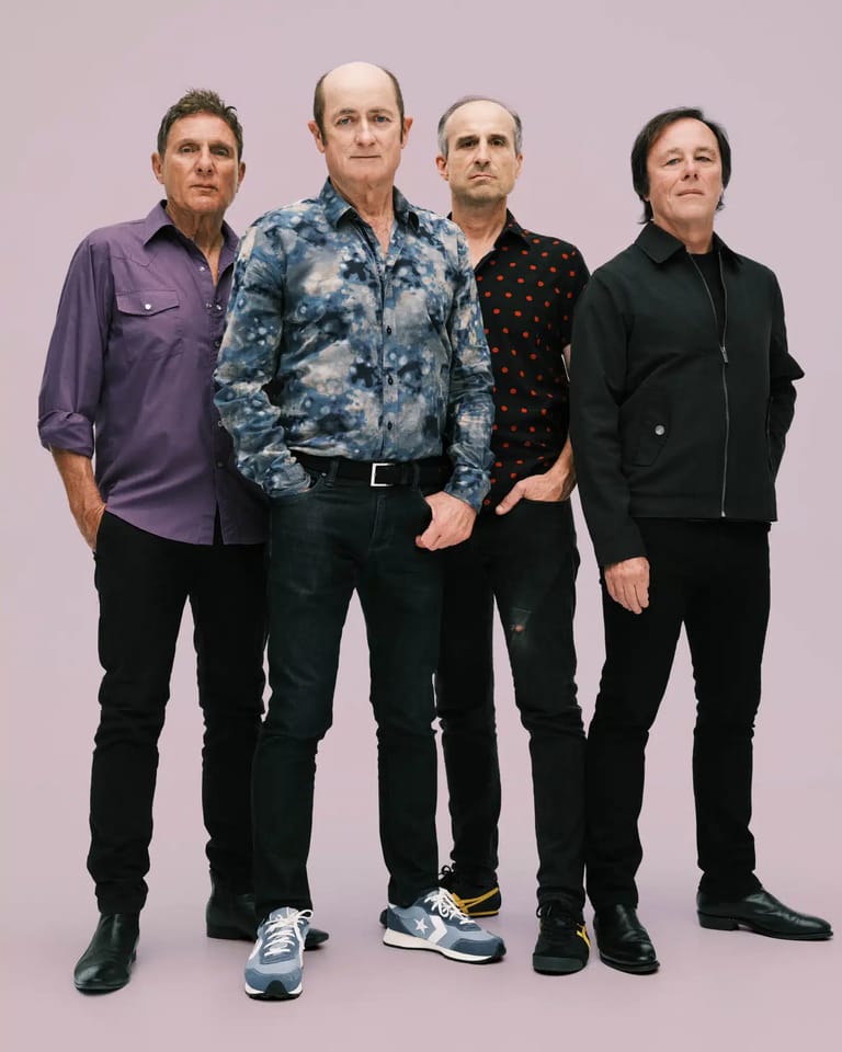 Four band members stand against a pale purple background, wearing casual shirts and dark trousers.