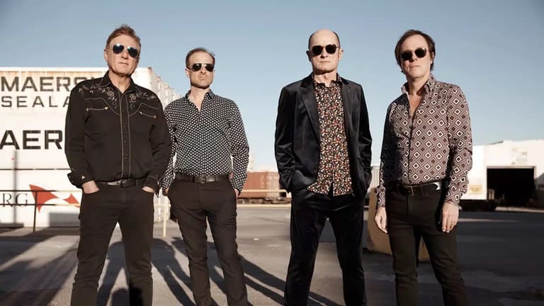 Four men in sunglasses stand shoulder-to-shoulder on a sunny rooftop or flat surface against an industrial backdrop.