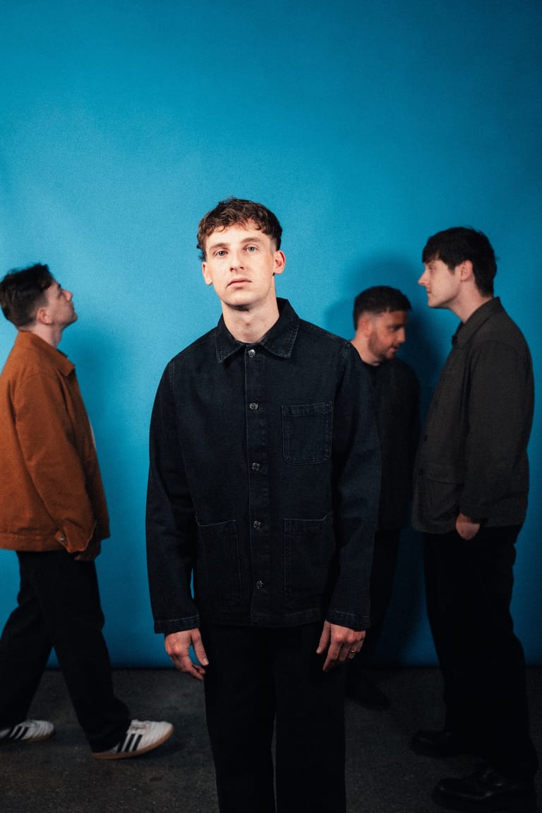 Four musicians stand against a bright blue backdrop, with one in focus wearing a dark shirt whilst others converse behind him.