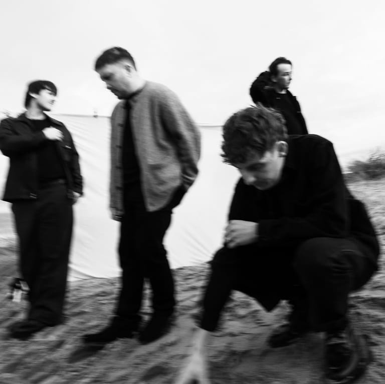 Four musicians stand together in a windswept outdoor landscape in this black and white photograph.