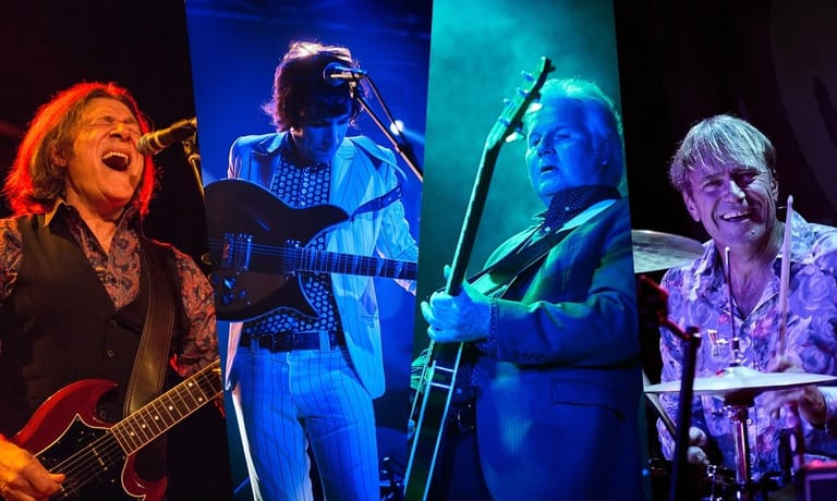 Four musicians perform on stage under coloured lighting, playing guitar, vocals, bass and drums during a live concert.