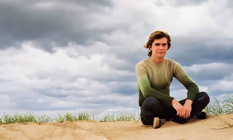 Young man in olive sweater crouched on sand against cloudy sky.