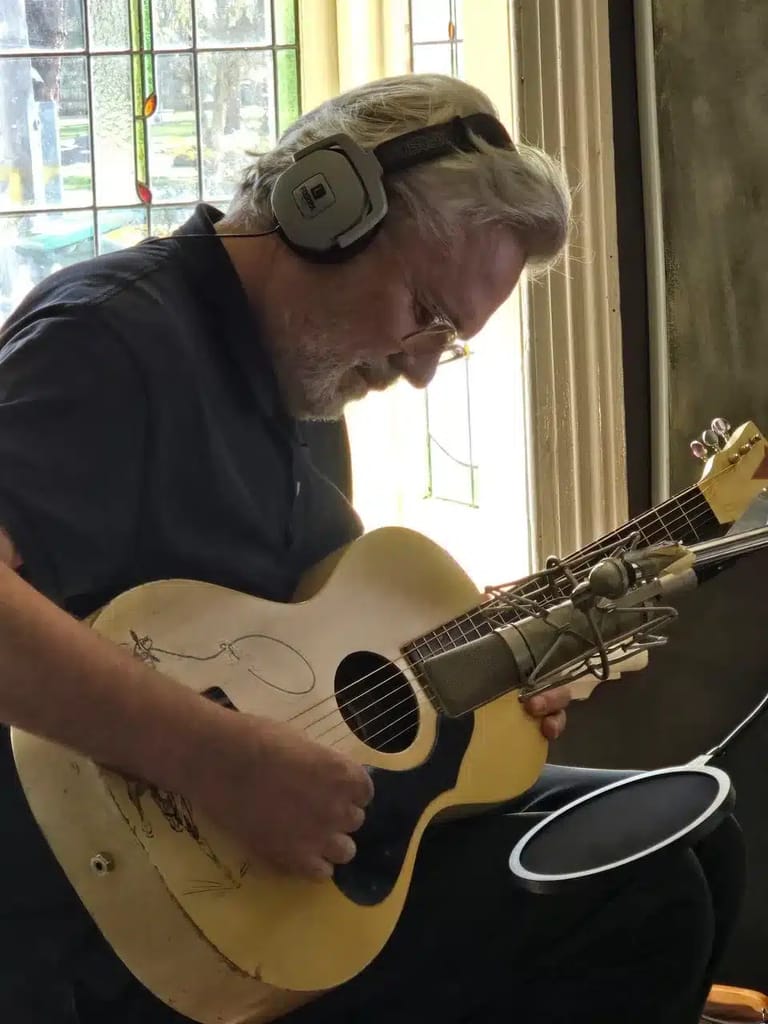 Musician wearing headphones plays an acoustic guitar by a sunlit window.