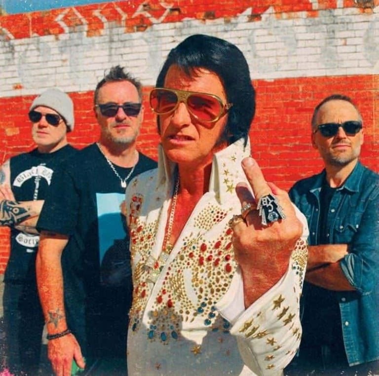 Four musicians in sunglasses pose against a red brick wall, the centre figure wearing a jewelled white and gold jumpsuit.