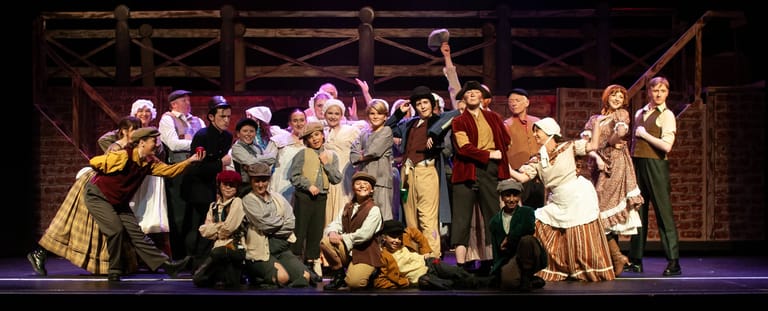 Large ensemble cast of young performers in period costume on a wooden stage set for Oliver!
