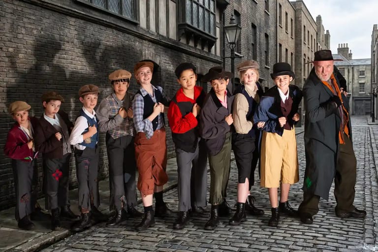 Cast of child and adult performers in period costume lined up on a cobblestone street for Oliver! stage production.