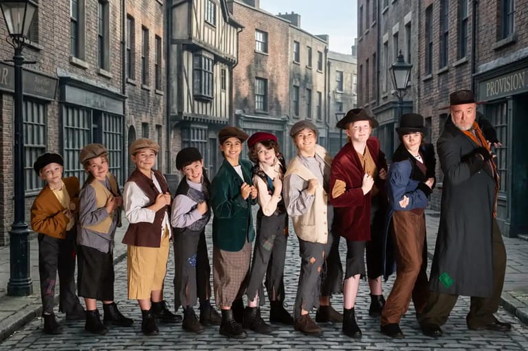 Cast of young performers and adults in Victorian-era costumes lined up on a period street set.