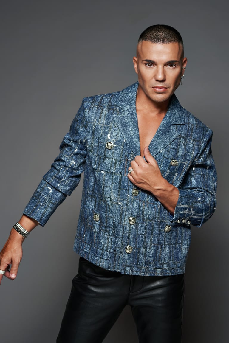 Man in blue textured blazer and black trousers poses against grey background.
