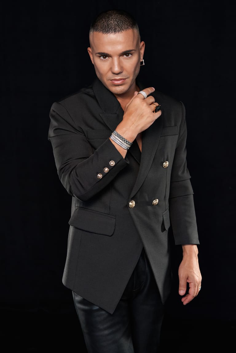 Person in black double-breasted blazer with hand raised to neck against dark background.