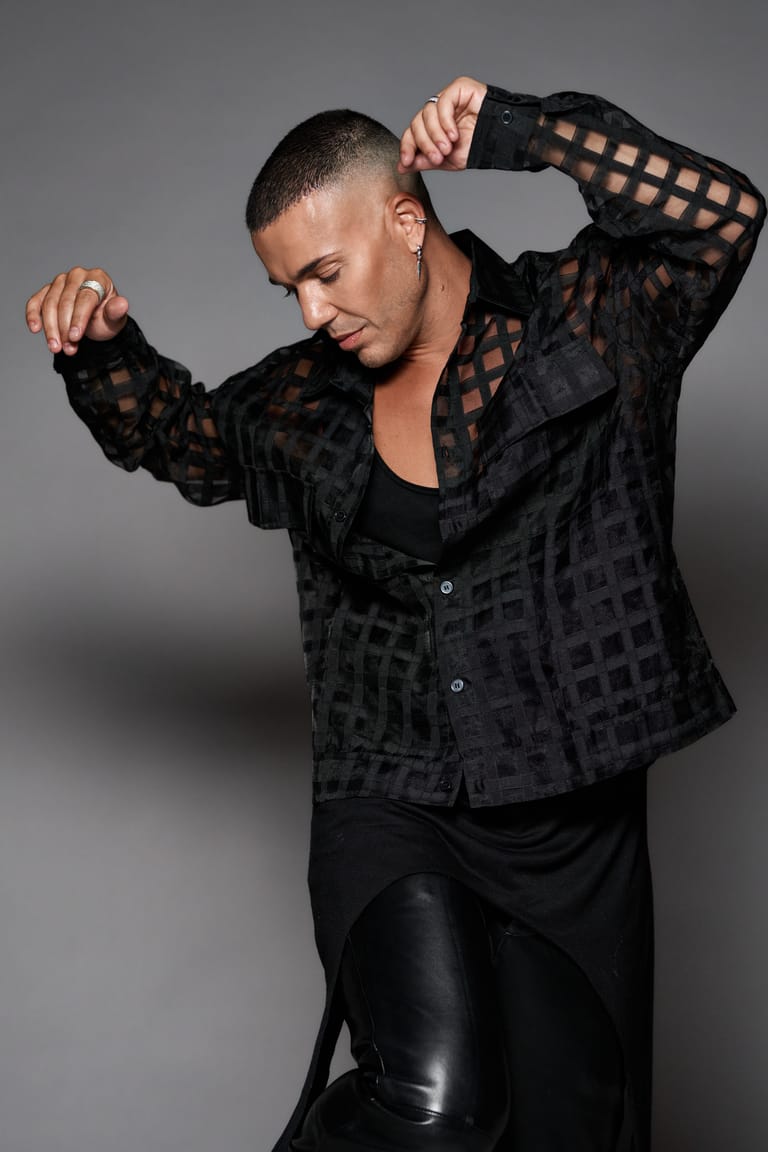 Performer in black patterned jacket and shiny trousers poses with hands raised against grey background.