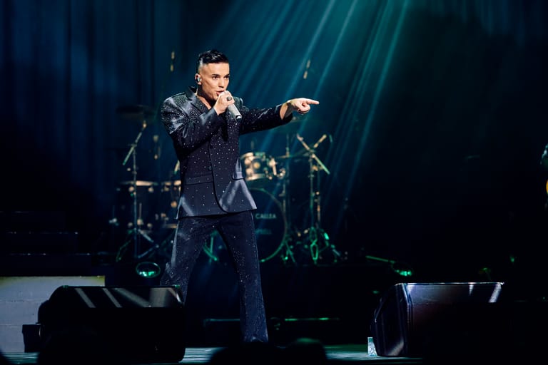 Performer in a dark suit pointing to the audience on stage with band members and dramatic blue lighting behind.