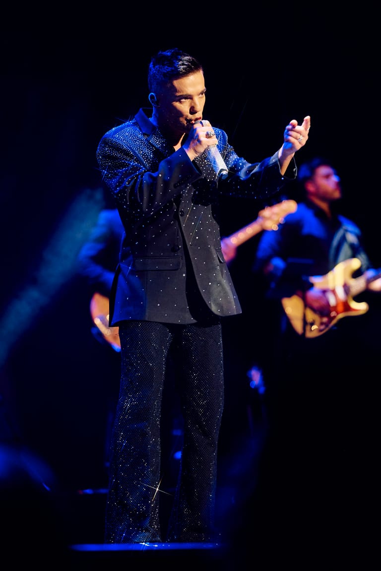 Performer in a sequined black suit singing into a microphone with band members playing behind on stage.