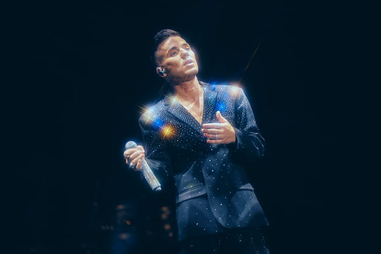 Performer in a sequined blue jacket singing under warm stage lighting during a tour performance.