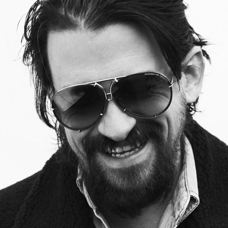 Man with dark hair and beard wearing aviator sunglasses, smiling at camera in black-and-white portrait.