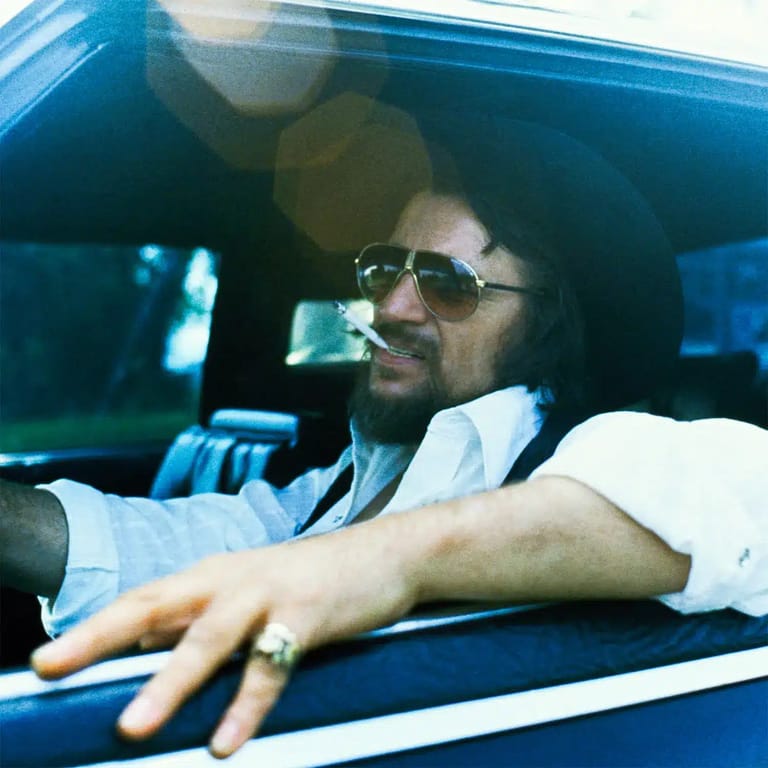 Man wearing sunglasses and light blue shirt seated in car with hand resting on window frame.