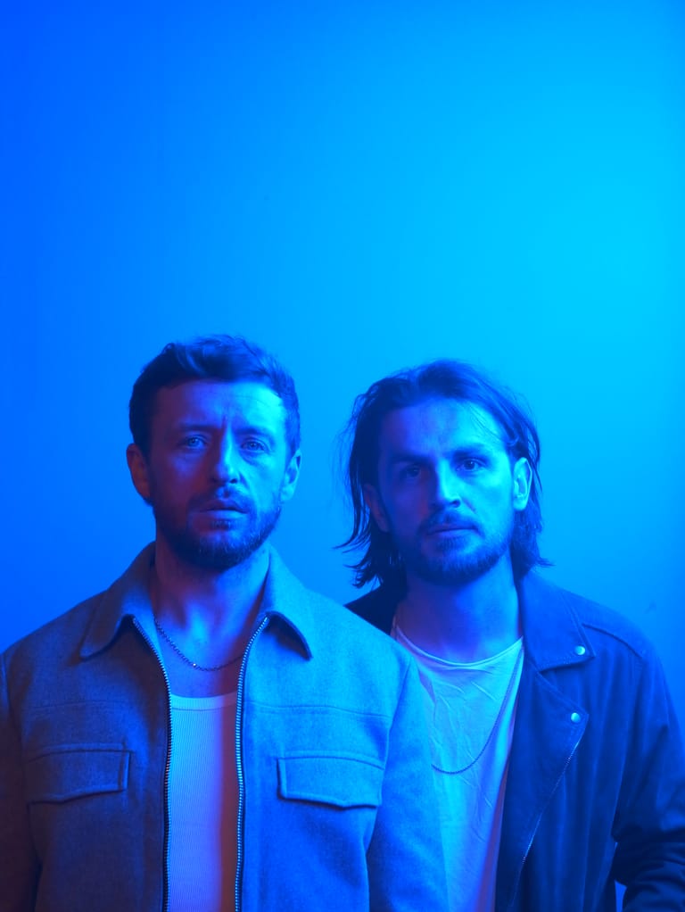 Two men stand against a blue-lit backdrop, wearing casual jackets and looking toward the camera with serious expressions.