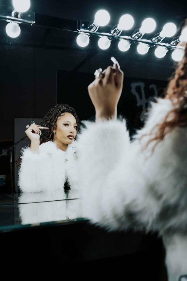 Performer applies makeup before a mirror lit by bright stage lights, wearing a white fluffy garment.