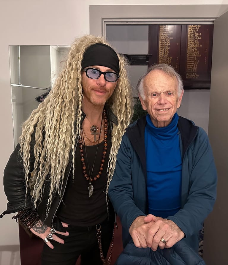 Two men pose together indoors, one wearing long blonde hair and black cap, the other in a blue jumper.