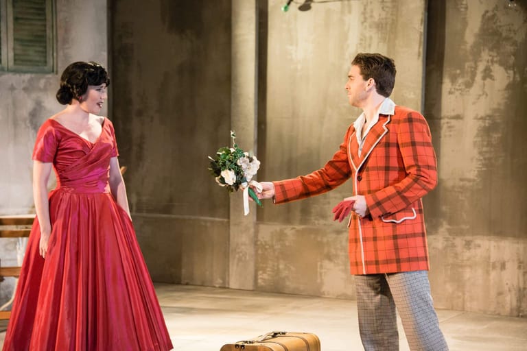 Man in orange plaid blazer presents flowers to woman in red dress on a theatre stage.