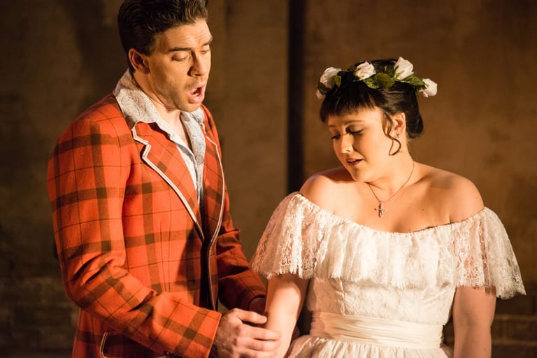 Two performers in period costume engage in a dramatic scene, one in an orange tartan jacket and the other in a white off-shoulder dress.