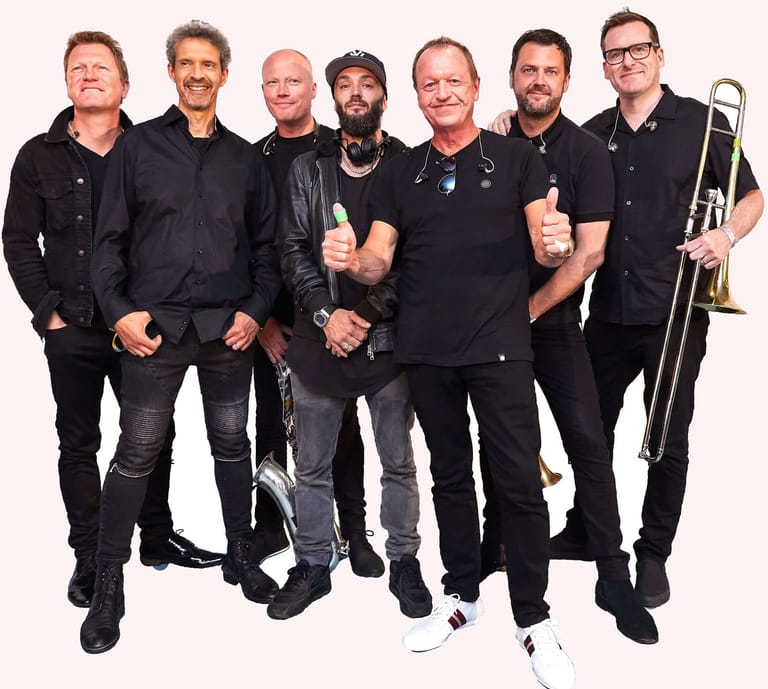 Seven male musicians in black clothing pose together with brass instruments, including trumpet and trombone.
