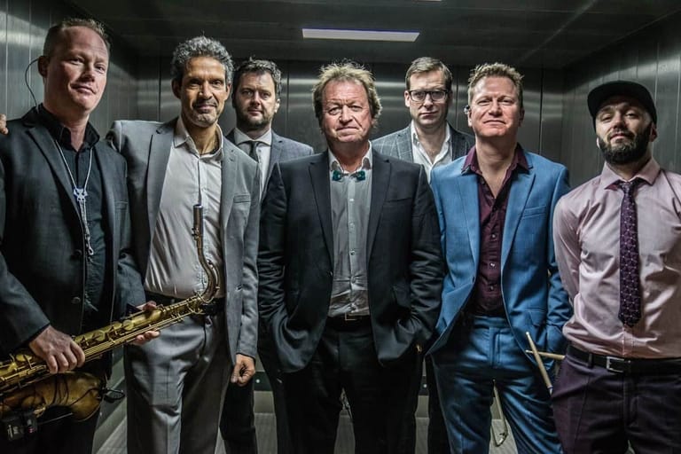 Seven musicians in suits stand in a line, with saxophonist on the left, against a grey interior backdrop.