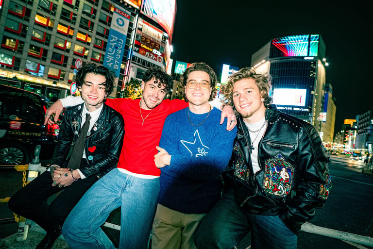 Four men in casual and leather jackets pose together on a neon-lit urban street at night.