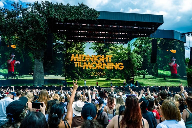 Crowd of festival-goers faces a large outdoor stage displaying "Midnight to Morning" branding under a metal canopy structure.